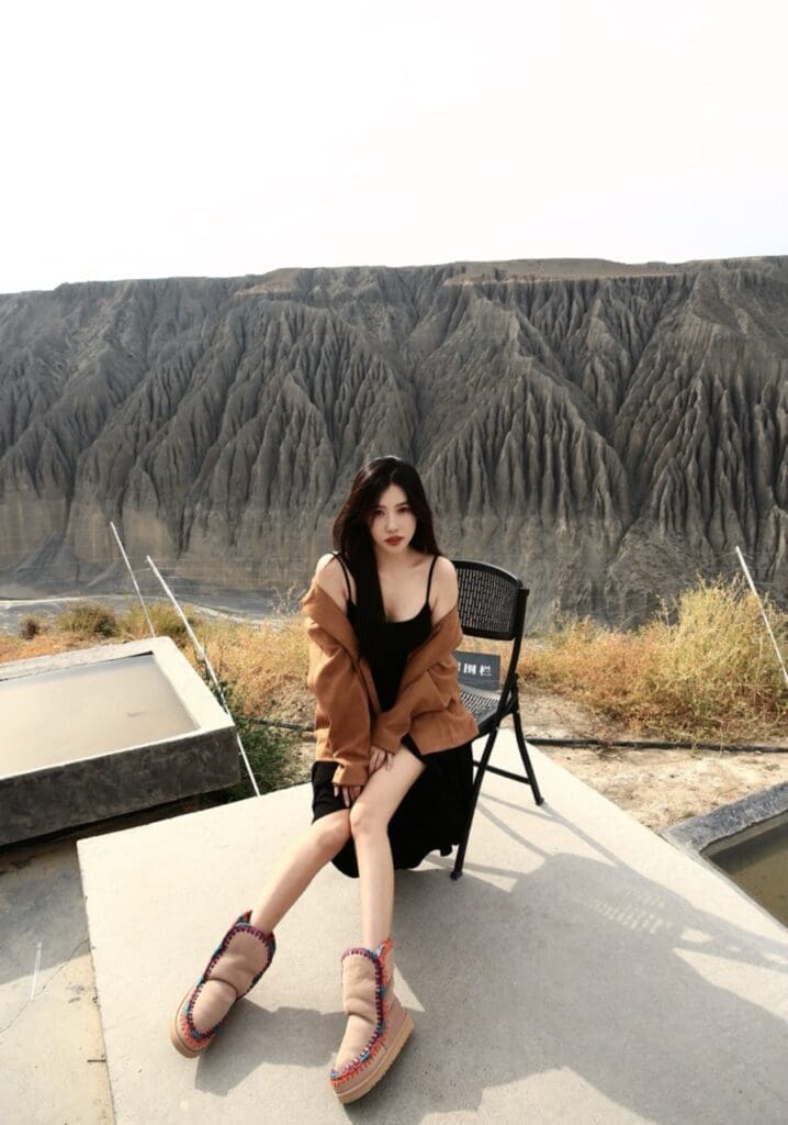 Chinese celebrity sitting on chair with mountain view behind her, wearing black dress, brown jacket, and brown mou shoes
